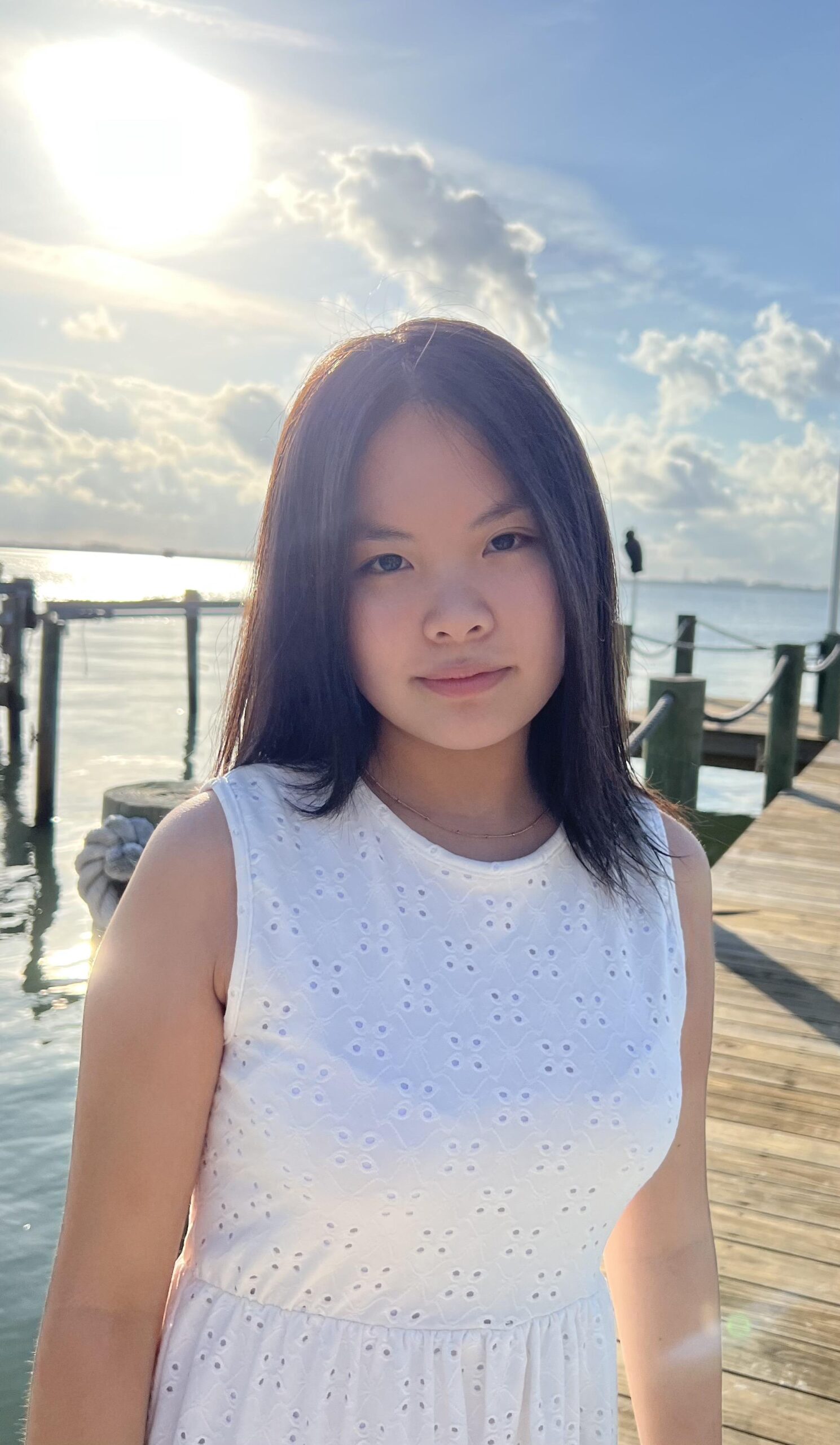 a girl in a white dress stands on a beach with the sun setting behind her