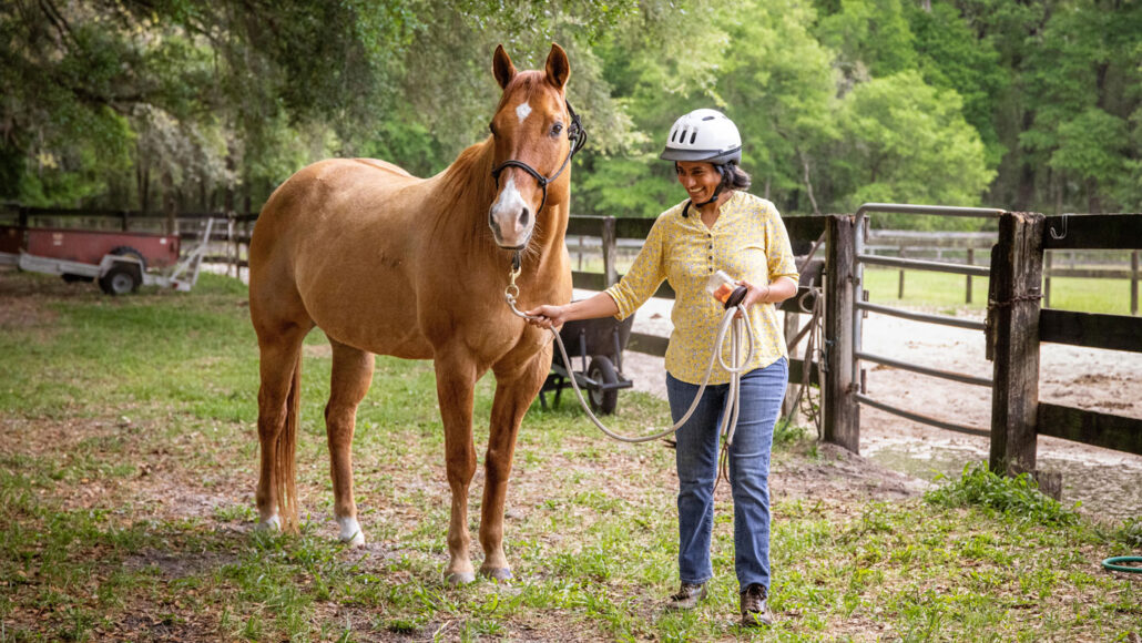 A photo of Eakta Jain standing next to a brown horse in a grassy area.