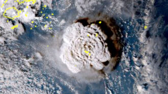 A satellite image of the Hunga Tonga volcano when it erupted in 2022 with a large circle of smoke visible in the middle of blue water and other clouds.