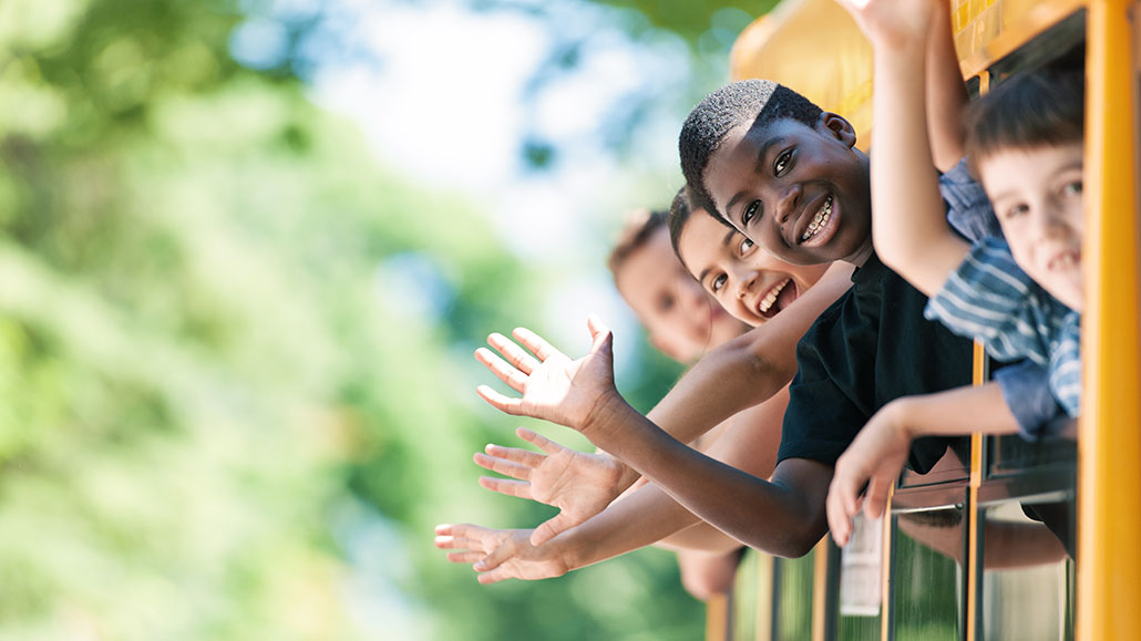 a bunch of kids are sticking their heads out of the windows of their yellow school bus. They are waving and smiling.