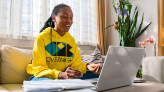 A Black woman wearing braids pulled back into a ponytail is smiling at her laptop. She is wearing a yellow hoody and she is sitting in a brightly lit living room.