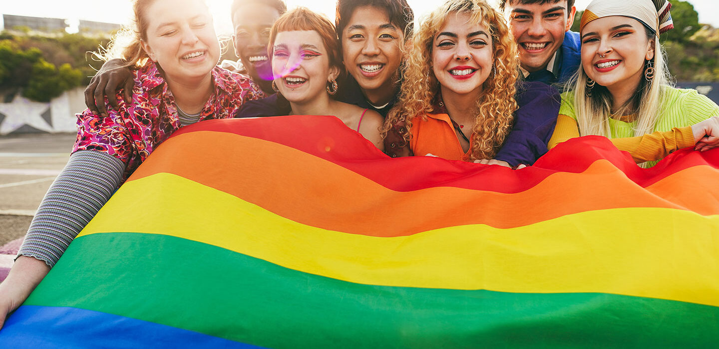 a photo showing a group of teens of different races and ages holding a Pride flag together