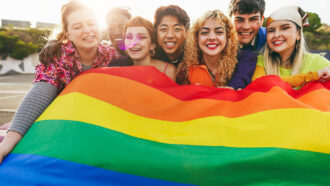 a photo showing a group of teens of different races and ages holding a Pride flag together