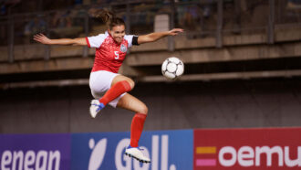 a woman in a white and red soccer uniform leaps into the air and draws back her foot to kick an incoming soccer ball