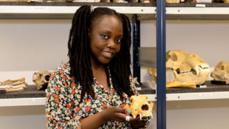 Pamela Akuku holds a skull and stands in front of a shelf full of fossils