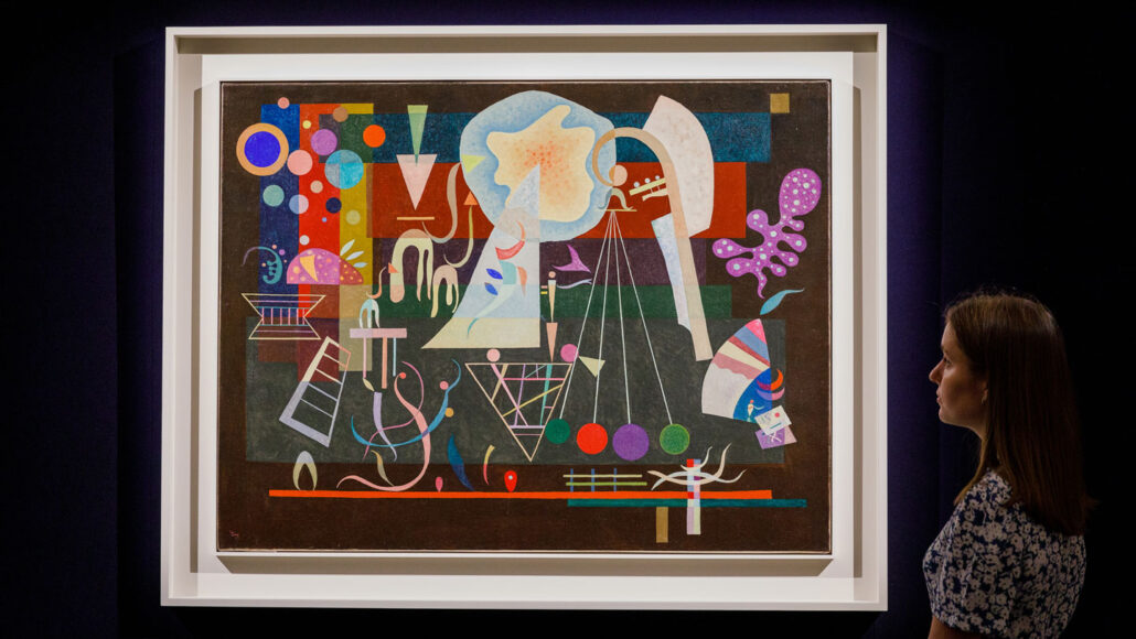an abstract painting with a medley of different shapes and colors hangs on a black wall; a woman in the corner of the frame gazes at the painting