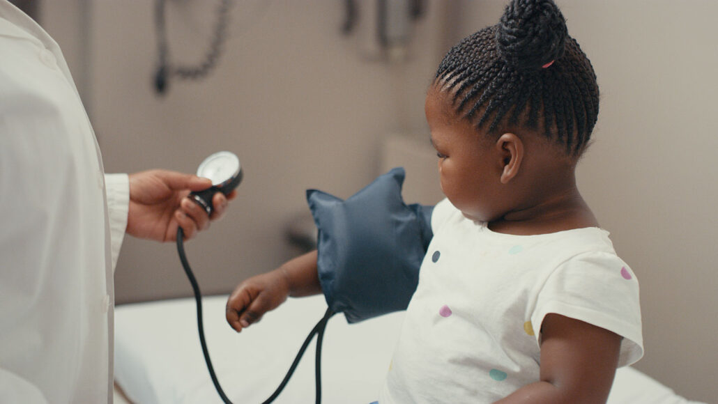 a toddler sits on the table of a doctor's office with a blood pressure cuff around her arm as the doctor takes a measurement
