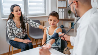 a tween in a tank top wears a blood pressure cuff around his upper arm while a doctor takes his blood pressure and his mother puts her hand on his shoulder