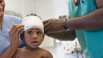 a young boy sits in his mother's lap while a doctor wearing scrubs wraps a white bandage around his head