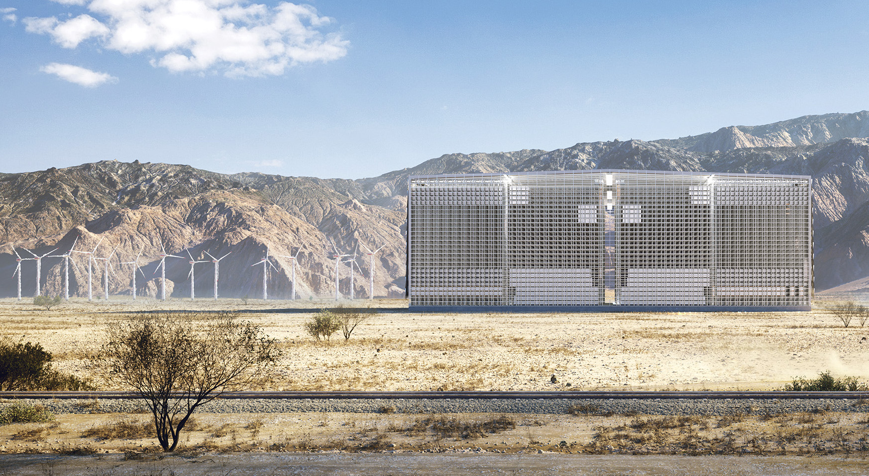 A large, silver building sits in the desert beside a fleet of wind turbines