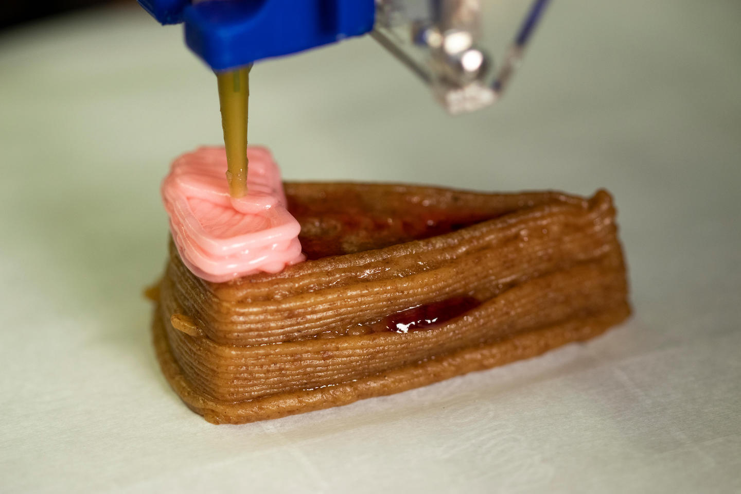 a cake-shaped wedge of 3-D printed brown batter is being topped with pink frosting by a 3-D printing nozzle