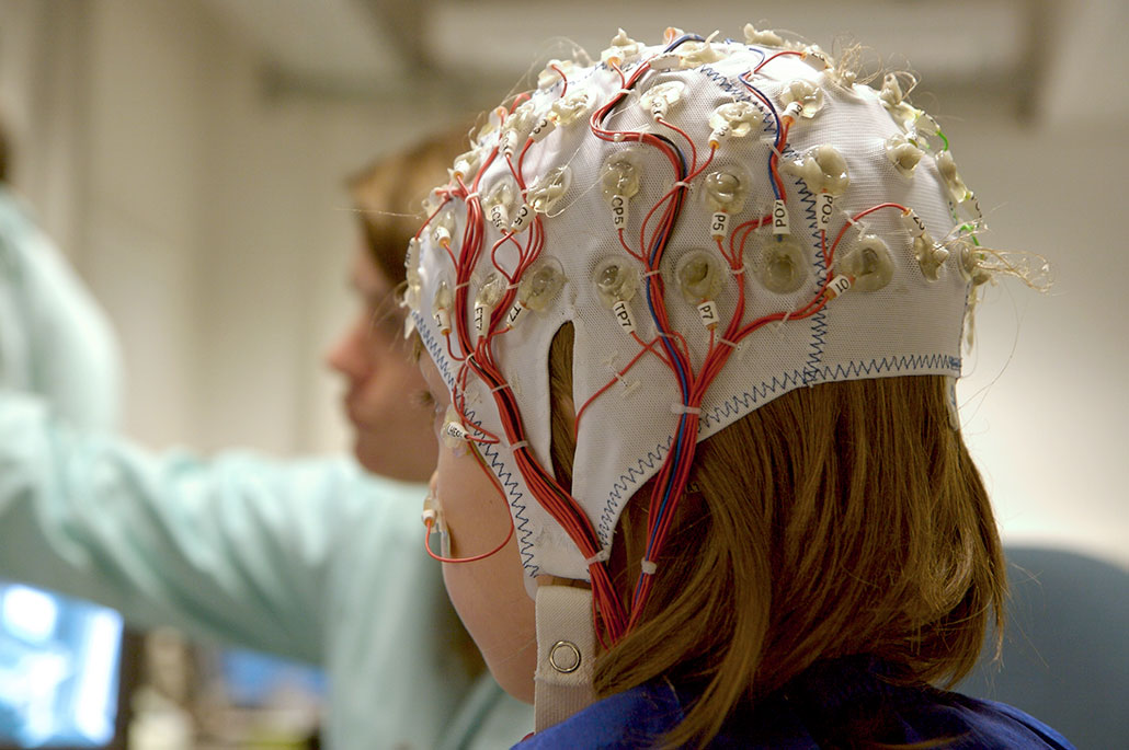 a girl wearing an EEG cap (a white cap over her hair with wireds and electrodes spread out like a net across the cap) and facing a blurry researcher in the background