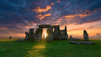 the stone circle of Stonehenge stands on a plain of green grass with the sun setting behind it