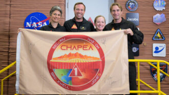 Four volunteers stand in front of the Mars Dune Alpha structure. They are holding up a mission flag and smiling