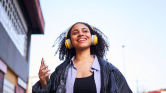 a teen girl in a leather jacket and bright yellow headphones smiles as she walks down the street holding a phone