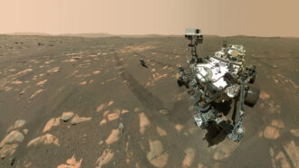 on rugged, reddish-brown terrain under a pinkish sky, a shiny metal rover stands beside a smaller drone-sized helicopter