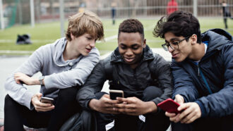 three guys sitting together outside near a soccer field, each has a phone but they are both looking at the phone of the person in the middle and smiling