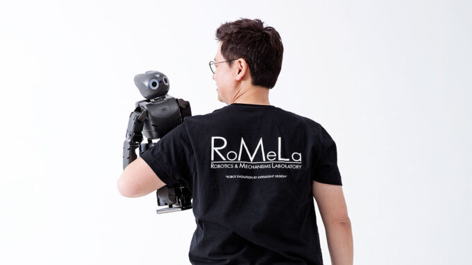 Dennis Hong stands facing away from the viewer in front of a white background. He is an Asian male with short black hair, and he is holding a robot in his left arm and looking at it.