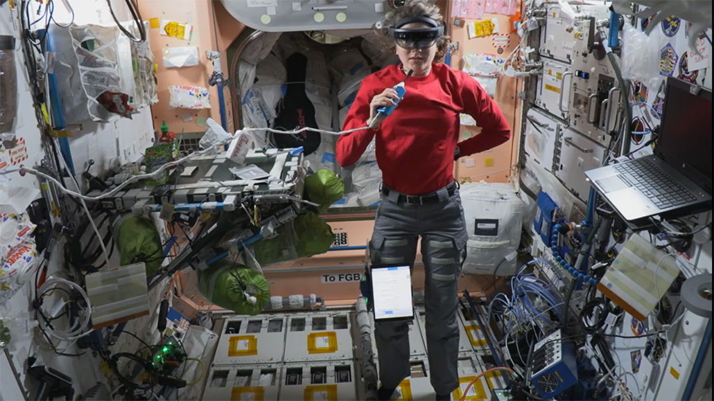a photo of Kayla Barron, an astronaut wearing a VR headset, a red long-sleeved shirt, and grey cargo pants, floating in the ISS module. She is speaking into a microphone