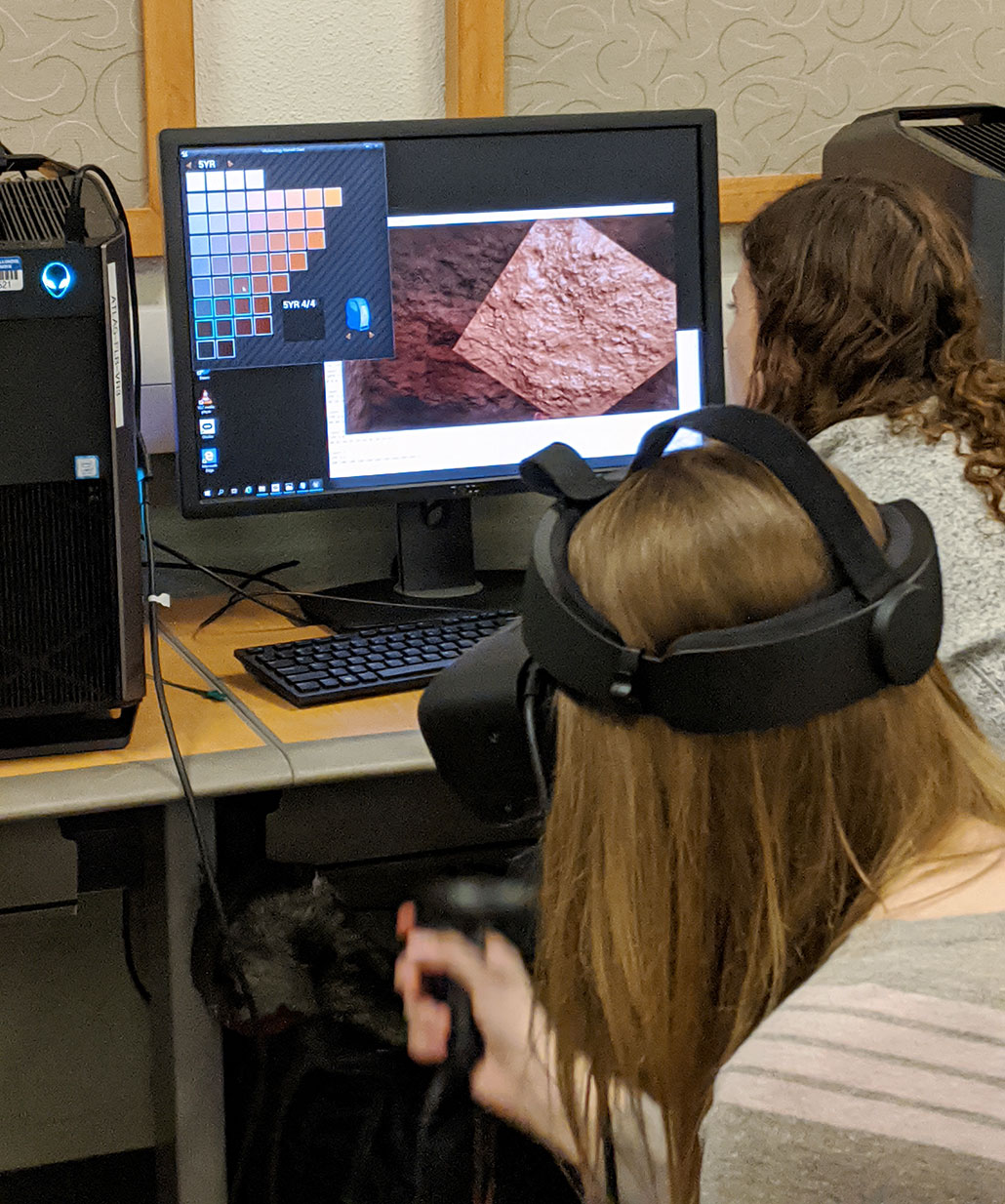 two students in front of a computer one is wearing a VR headset and using a hand controller, the other is looking at the monitor. The monitor is showing a cave surface texture