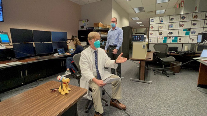 a photo of an older white man sitting in a chair in a mostly empty office. His left hand is outreached to "shake hands" with the astronaut he is speaking with.