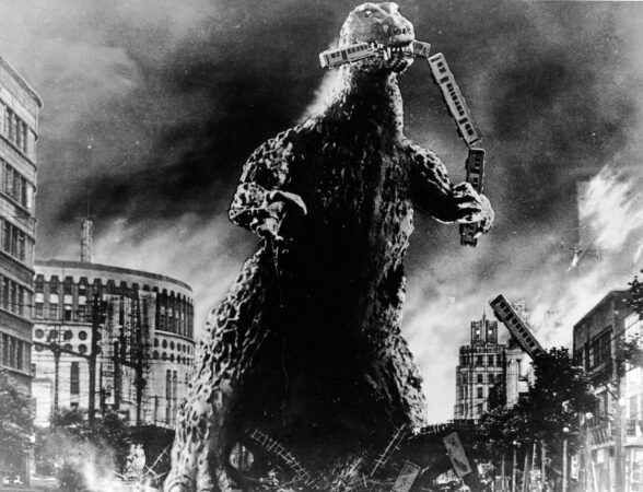 Godzilla holds a commuter train in his mouth. A ruined city surrounds him.