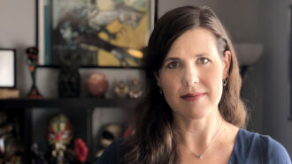 Sociologist Margee Kerr stands in front of a shelf lined with sculptures. Kerr has long, wavy brown hair and hazel eyes. She is wearing a blue shirt and a necklace.