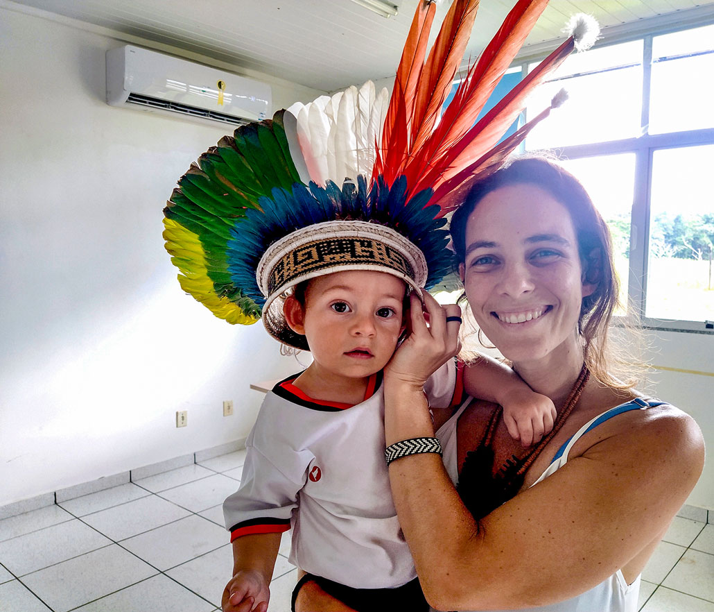 Carolina Levis holds her son, Davi. He is wearing a headdress with colorful feathers. They are standing in a white room.