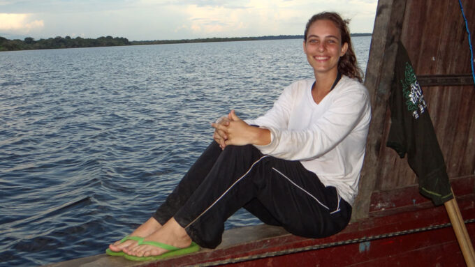 Ecologist Carolina Levis sits on a boat. A calm Lake Acara is behind her. She has long brown hair and is wearing a white long sleeved shirt.