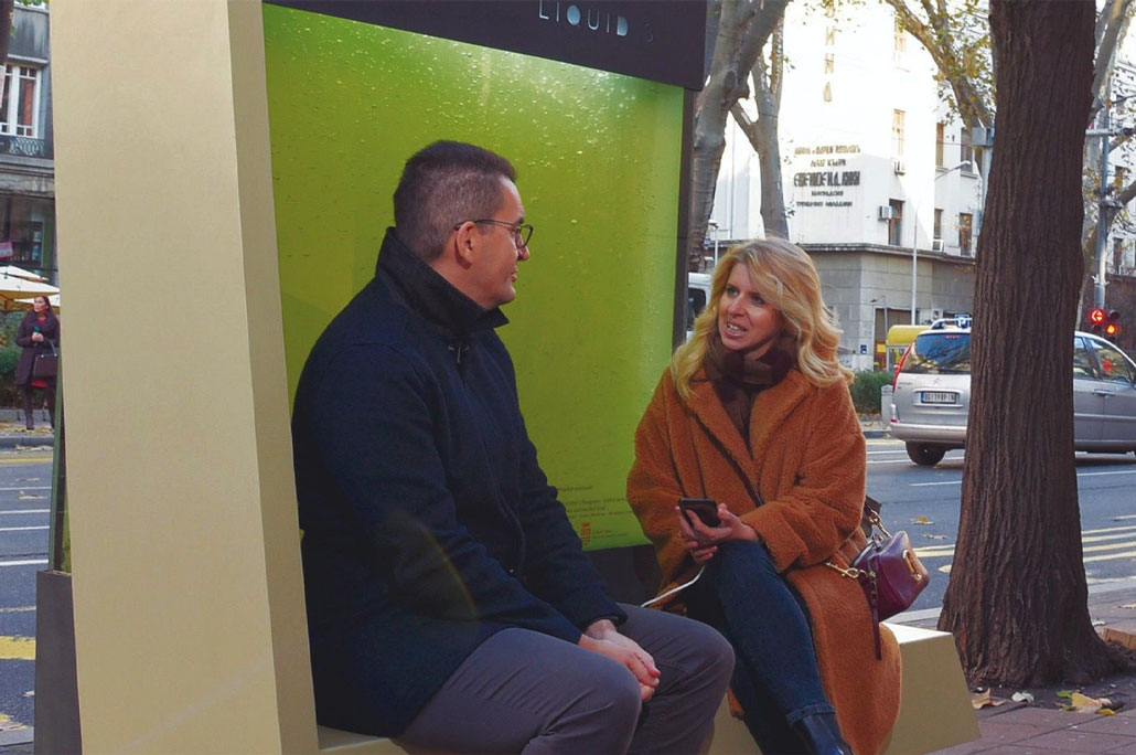 2 people sitting on a bench with a tank of green algae behind them