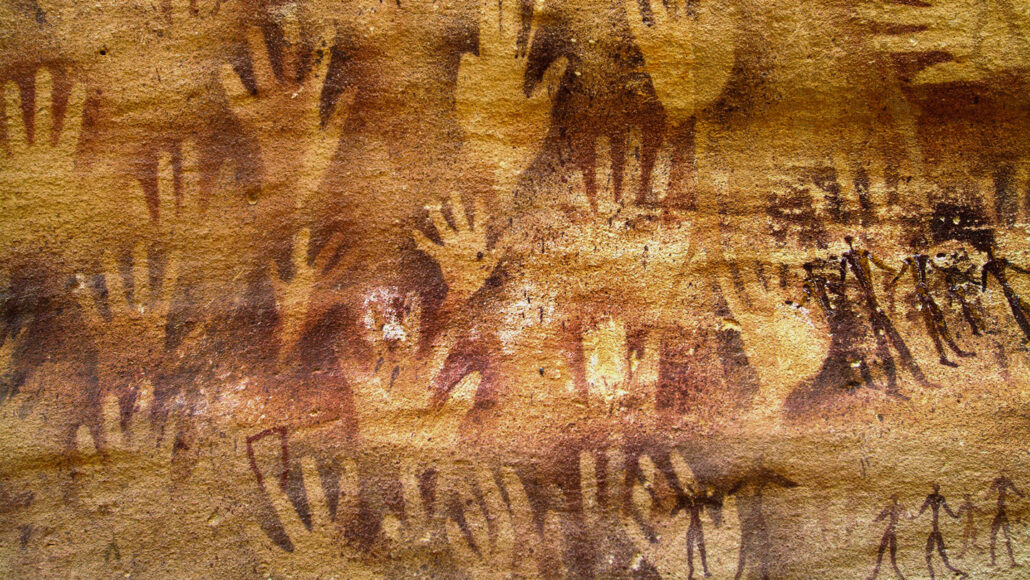 the reddish-brown wall of a cave is imprinted with human hand shapes and decorated with cave paintings of stick figures