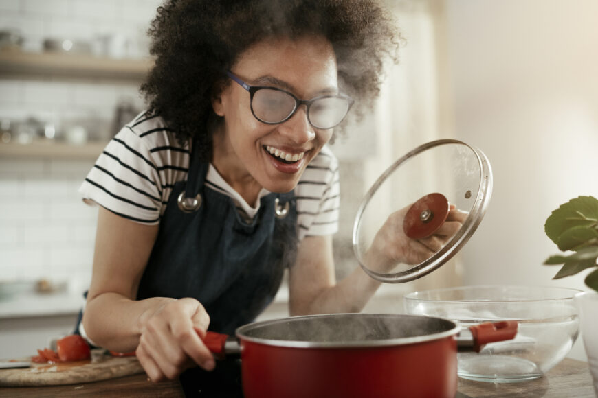 A young woman with curly hair and steamed glasses is opening the lid of a red pot.