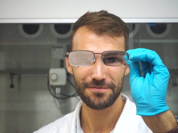 Chemist Alexander Henn holds up a pair of glasses with one gloved hand. The right lens is fogged up while the left lens is clear due to the coating.