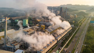 an aerial view of a factory beside a highway shows plumes of air pollution drifting from smoke stacks