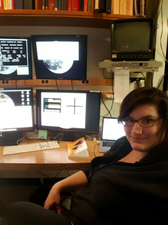 Astrophycist is sitting at a desk in her control room. She is in front of four computer monitors. A laptop is next to her right arm.