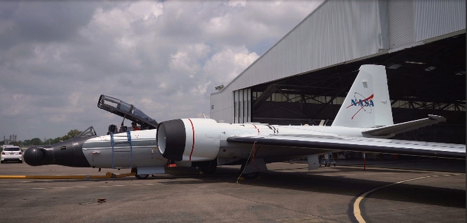 A small, two-seater jet with a NASA logo on its tail sits just outside a hangar on a cloudy day.