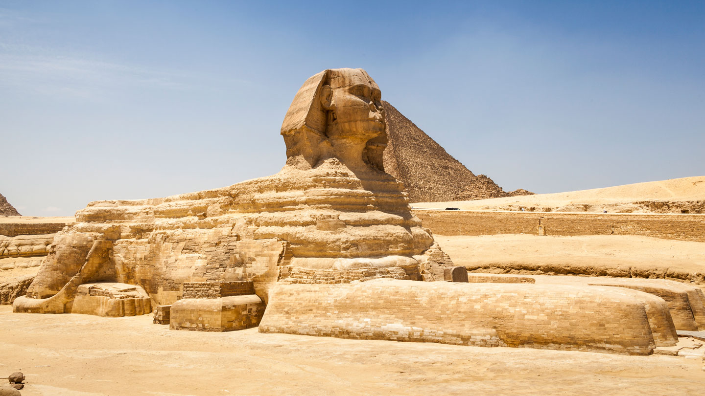 A huge earthen formation in the shape of a sphinx stands in front of the Great Pyramid of Giza under a blue sky