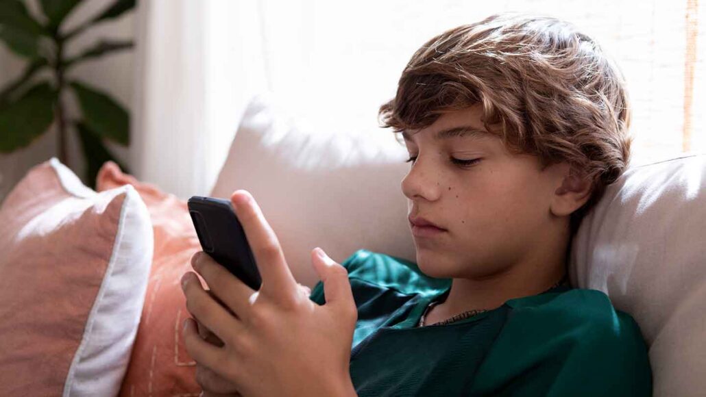 A young teen with short blond hair is wearing a green shirt and sitting on a sofa. He is looking at his smartphone.