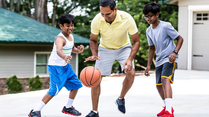 A father and two kids playing basketball on their driveway outside their house. The ball is mid-bounce.