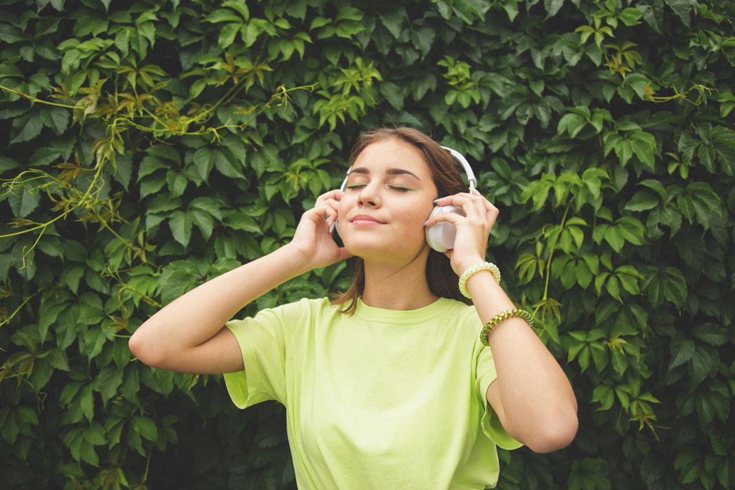 a young woman with pale skin and brown hair listening to music on headphones. She is in front of a leafy background and has a green shirt and matching green eyeliner.