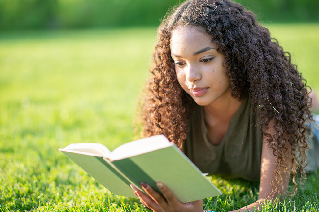 a girl with brown skin and curly hair is laying in a field of grass and reading