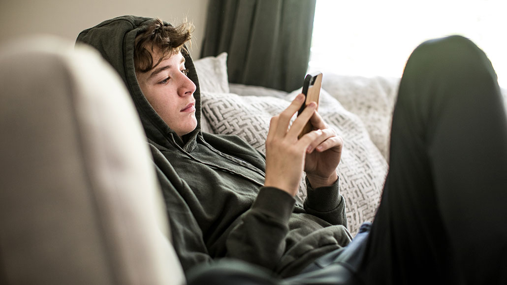 a photo of a kid wearing an olive green sweatsuit with the hood up. He has dark brown curly hair and pale skin and is scrolling on his smartphone.