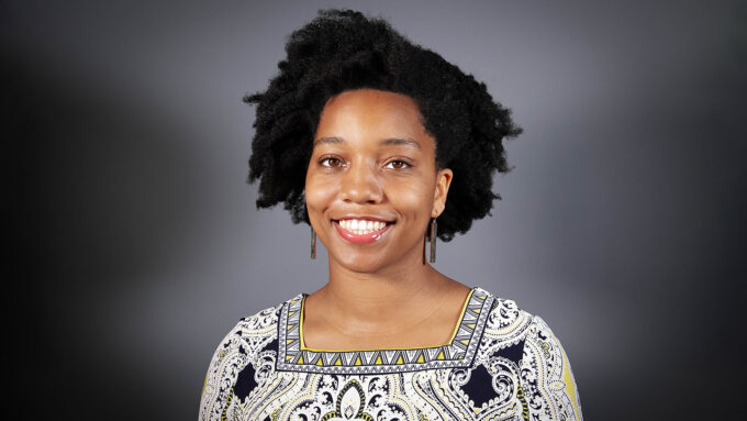 Civil Engineer Destenie Nock stands in front of a gray starburst background. She is a black woman with medium length, curly hair. She is wearing a tan blouse with black swirls and patterns.
