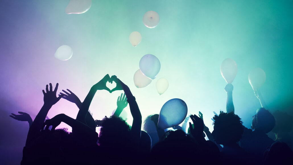 a crowd of people dancing with hands in the air as ballons fall, the lights in front of the crowd are a teal to purple gradient