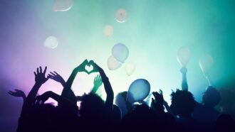a crowd of people dancing with hands in the air as ballons fall, the lights in front of the crowd are a teal to purple gradient