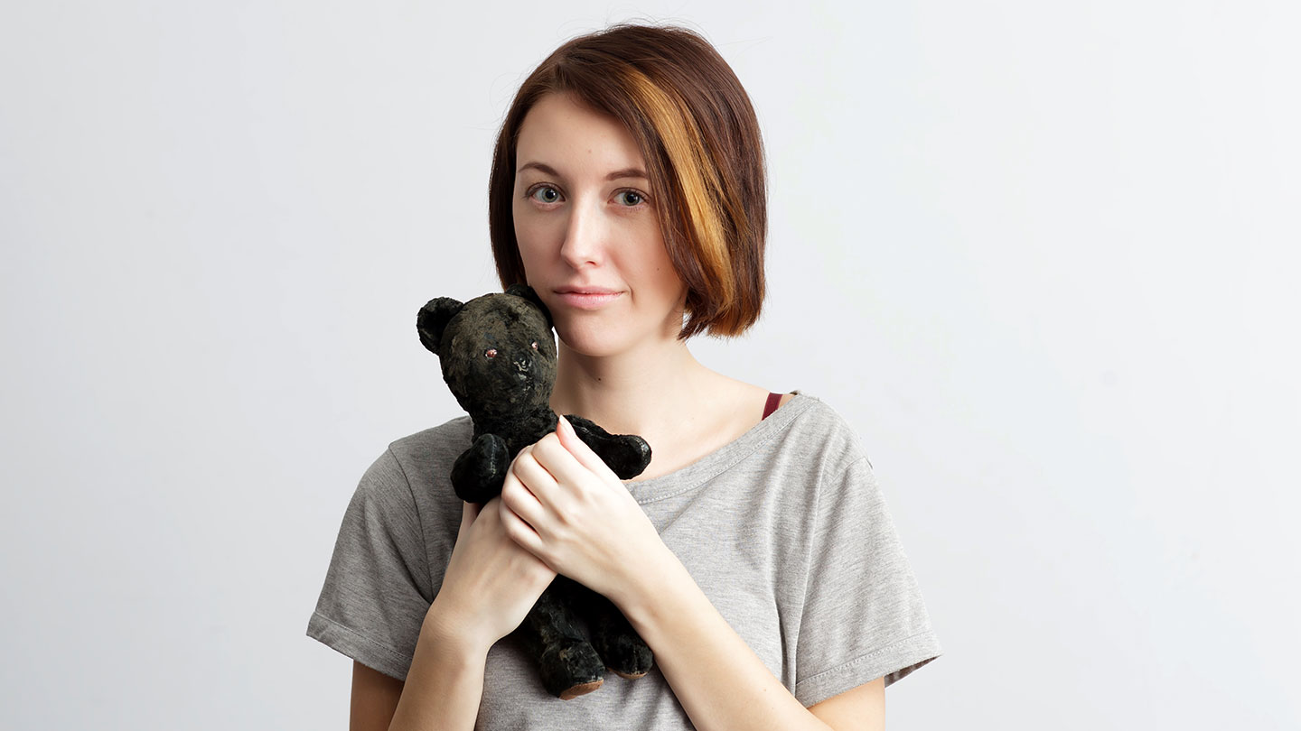 a photo of a young woman with pale skin and short brown hair holding a very well-loved (worn out) teddy bear