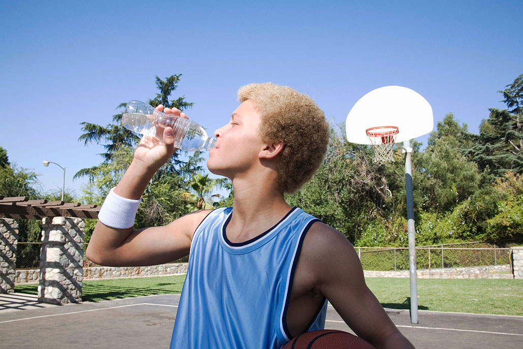 a young man with a light brown to blond afro drinking from a water bottle. He is standing on a basketball court and holding a basketball in one arm