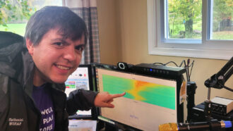 Space physicist Nathaniel Frissell smiles at the camera. He is a white man with brown eyes and brown hair. He is pointing to a computer screen with a colorful chart on it.