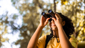 a smiling girl surrounded by trees holds a set of binoculars up to her eyes to peer upward