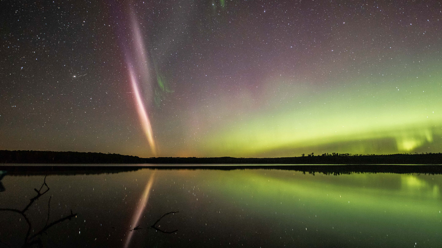 a photo of STEVE and the green picket fence skyglows in a starry night sky. Still water refects the sky. On the far right is a typical green aurora.